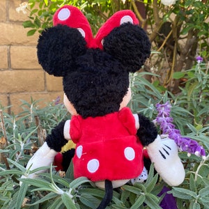 Pu&ograve; includere: Una bambola di peluche di Minnie Mouse, vista da dietro, con un vestito e un fiocco rossi, entrambi adornati da pois bianchi. La bambola ha i capelli neri e guanti bianchi. Lo sfondo presenta fogliame verde e un muro di mattoni.