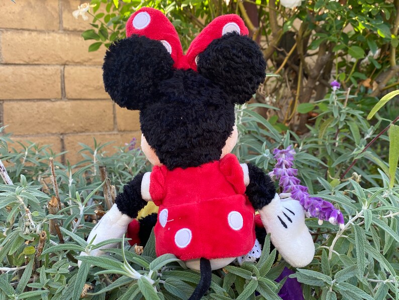 Pu&ograve; includere: Una bambola di peluche di Minnie Mouse, vista da dietro, con un vestito e un fiocco rossi, entrambi adornati da pois bianchi. La bambola ha i capelli neri e guanti bianchi. Lo sfondo presenta fogliame verde e un muro di mattoni.