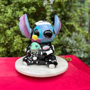 May include: A ceramic figurine of a blue alien-like character with large ears, holding a green creature. The figurine is painted with black and white bone-like patterns and sits on a white marble-like base. The background is blurred green foliage.