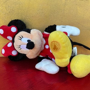 Pu&ograve; includere: Un peluche di Minnie Mouse, sdraiata. Minnie ha testa e orecchie nere, un fiocco rosso a pois bianchi e un vestito rosso. Il suo piede giallo ha un emblema dorato. Sono visibili guanti e scarpe bianche.