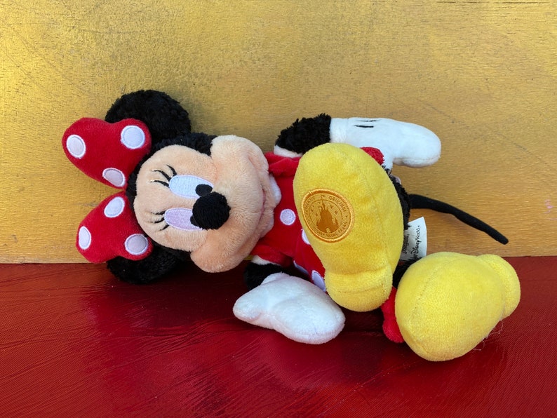 Pu&ograve; includere: Un peluche di Minnie Mouse, sdraiata. Minnie ha testa e orecchie nere, un fiocco rosso a pois bianchi e un vestito rosso. Il suo piede giallo ha un emblema dorato. Sono visibili guanti e scarpe bianche.