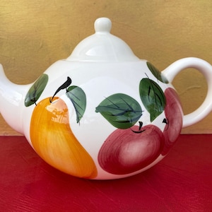 May include: White ceramic teapot with a lid, spout, and handle. The teapot is decorated with hand-painted fruit, including a large yellow pear and red apples with green leaves. The teapot sits on a red surface.