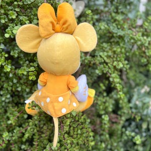 Pode incluir: Um boneco de pel&uacute;cia de rato amarelo com um grande la&ccedil;o na cabe&ccedil;a, vestindo um vestido amarelo com bolinhas brancas. O boneco tem orelhas grandes e est&aacute; colocado em um fundo de folhagem verde.