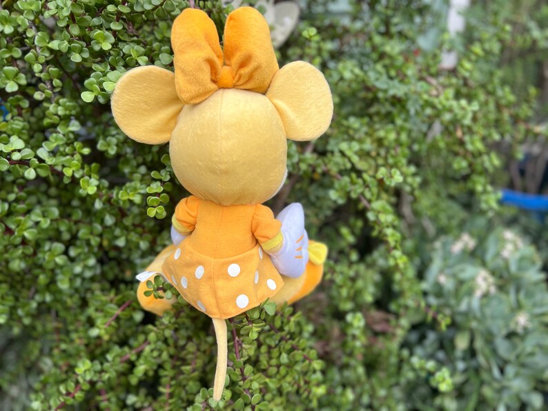 Pode incluir: Um boneco de pel&uacute;cia de rato amarelo com um grande la&ccedil;o na cabe&ccedil;a, vestindo um vestido amarelo com bolinhas brancas. O boneco tem orelhas grandes e est&aacute; colocado em um fundo de folhagem verde.
