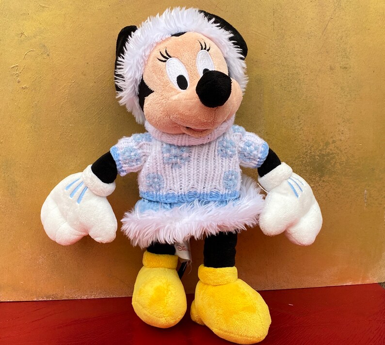 K&ouml;nnte beinhalten: Ein Pl&uuml;schtier von Mickey Mouse's Freundin Minnie Mouse, die einen wei&szlig;en und blauen Strickpullover mit Schneeflockenmuster, einen wei&szlig;en Pelzbesatz und einen wei&szlig;en Hut mit blauem Besatz tr&auml;gt.