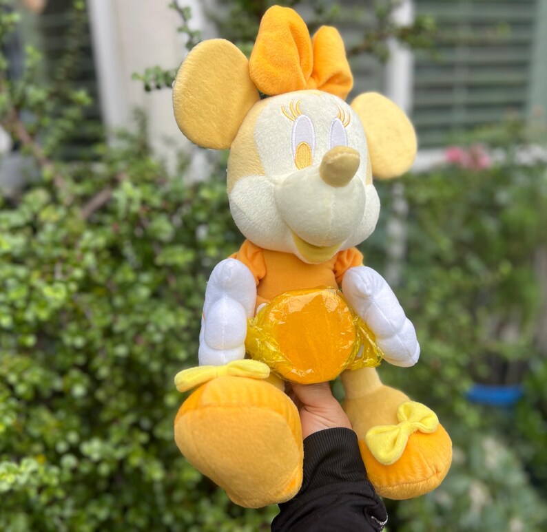 Pode incluir: Uma boneca de pel&uacute;cia da Minnie Mouse em tons de amarelo e branco. A boneca tem um la&ccedil;o, orelhas e sapatos amarelos com la&ccedil;os. Ela usa um vestido amarelo e luvas brancas. A boneca &eacute; macia e fofinha.