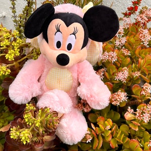 May include: Pink plush toy of a cartoon mouse with large black ears and yellow and white checkered accents. The toy is sitting in a bed of green and brown plants with small pink flowers.