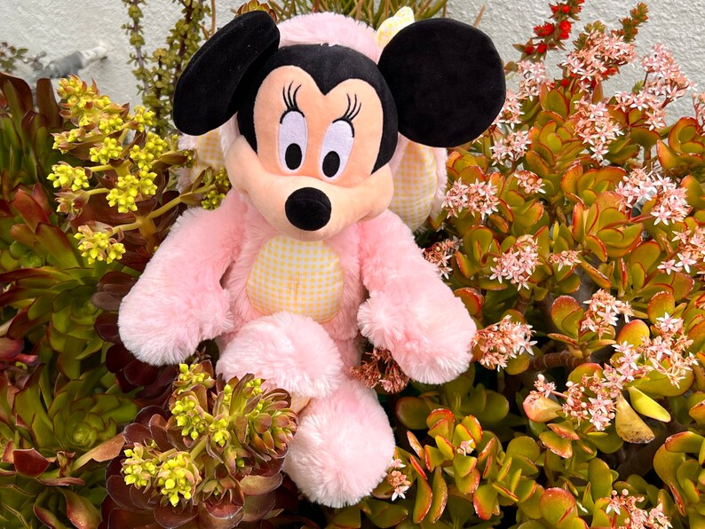 May include: Pink plush toy of a cartoon mouse with large black ears and yellow and white checkered accents. The toy is sitting in a bed of green and brown plants with small pink flowers.