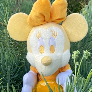 Pode incluir: Uma boneca de pel&uacute;cia da Minnie Mouse com la&ccedil;o, orelhas e vestido amarelos. A boneca tem um rosto branco com tra&ccedil;os bordados e &eacute; cercada por folhagens verdes. A boneca tem cerca de 20 cm de altura.