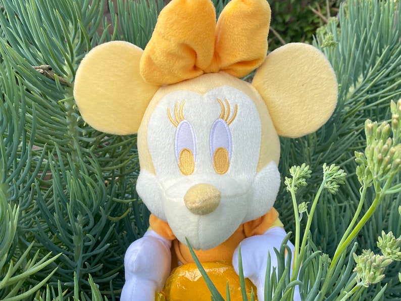 Pode incluir: Uma boneca de pel&uacute;cia da Minnie Mouse com la&ccedil;o, orelhas e vestido amarelos. A boneca tem um rosto branco com tra&ccedil;os bordados e &eacute; cercada por folhagens verdes. A boneca tem cerca de 20 cm de altura.