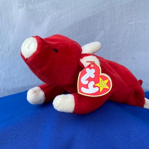 May include: A red plush toy bull with white accents on its snout, feet, and horns. The toy has a tag attached with the words "Beanie Baby" on it. The toy is lying on a blue surface.