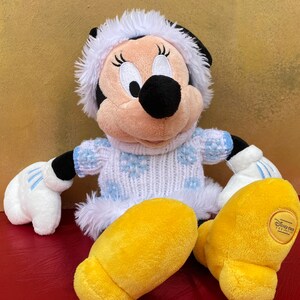 K&ouml;nnte beinhalten: Ein Pl&uuml;schtier von Minnie Mouse, das einen wei&szlig;en und blauen Pullover mit Schneeflocken, einen wei&szlig;en, mit Fell besetzten Hut und gelbe Schuhe tr&auml;gt. Das Spielzeug hat ein Disney Parks-Etikett am Fu&szlig;.