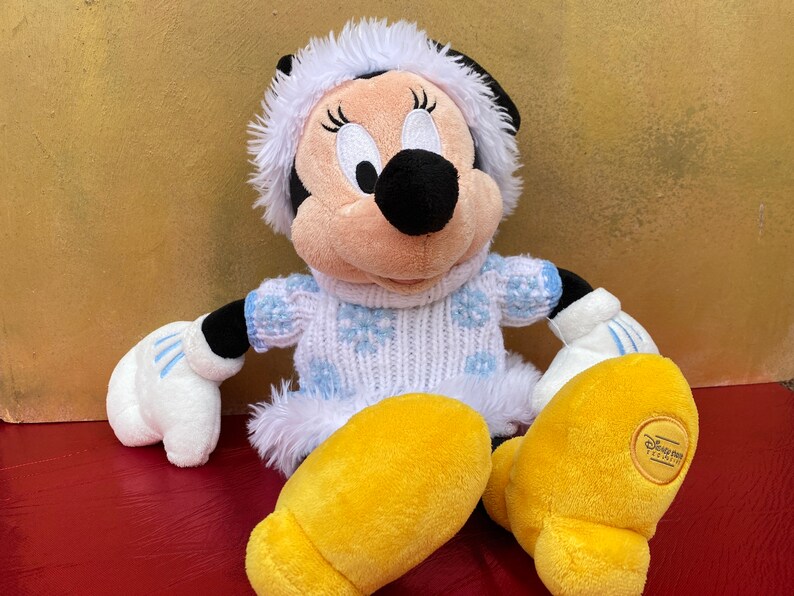 K&ouml;nnte beinhalten: Ein Pl&uuml;schtier von Minnie Mouse, das einen wei&szlig;en und blauen Pullover mit Schneeflocken, einen wei&szlig;en, mit Fell besetzten Hut und gelbe Schuhe tr&auml;gt. Das Spielzeug hat ein Disney Parks-Etikett am Fu&szlig;.