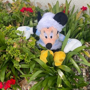K&ouml;nnte beinhalten: Ein Pl&uuml;schtier eines Comic-Charakters, das einen wei&szlig;en Hut und blaue und wei&szlig;e Kleidung tr&auml;gt. Das Spielzeug sitzt in einem Bett aus gr&uuml;nen Pflanzen mit roten Blumen.