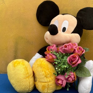 May include: A plush Mickey Mouse doll with black ears, a black body, and yellow shorts. The doll is holding a bouquet of pink and orange roses with green leaves. The background is a solid yellow color.
