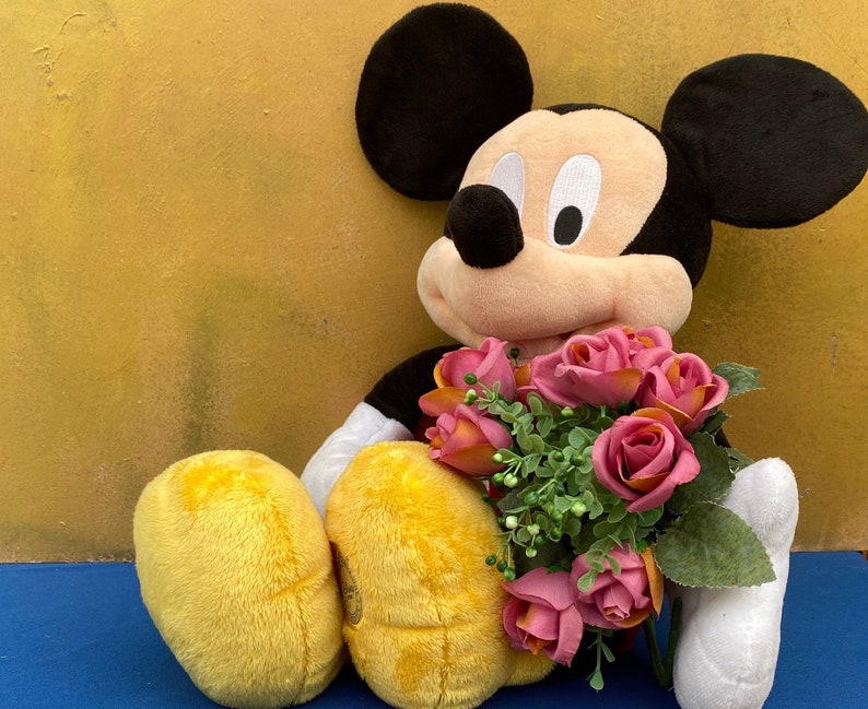 May include: A plush Mickey Mouse doll with black ears, a black body, and yellow shorts. The doll is holding a bouquet of pink and orange roses with green leaves. The background is a solid yellow color.