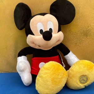 May include: A plush Mickey Mouse doll, featuring black ears, a black body, red shorts, and yellow feet. The doll has white gloves and a smiling face. A tag is attached to the doll.