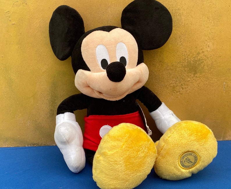 May include: A plush Mickey Mouse doll, featuring black ears, a black body, red shorts, and yellow feet. The doll has white gloves and a smiling face. A tag is attached to the doll.