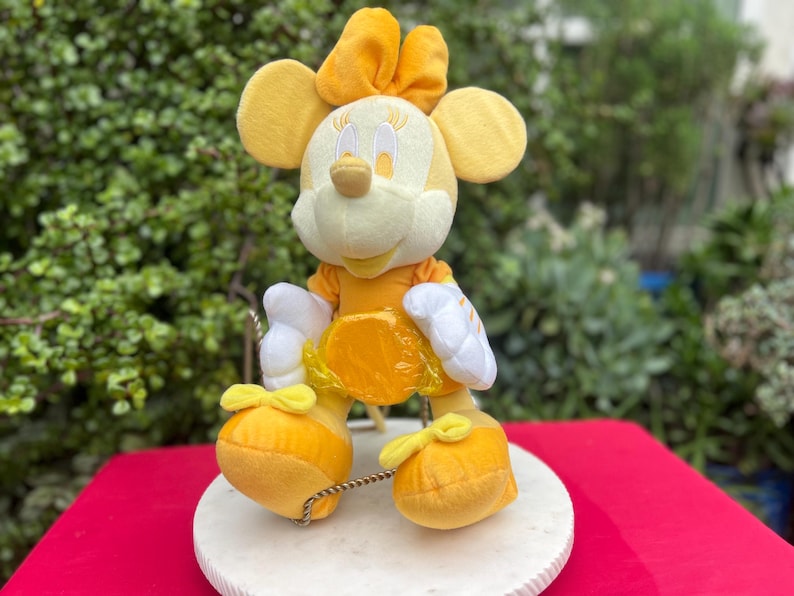 Pode incluir: Uma figura de pel&uacute;cia da Minnie Mouse em tons de amarelo e branco. A figura tem orelhas grandes e redondas, um la&ccedil;o e um vestido amarelo. Est&aacute; em uma base redonda branca, com um fundo vermelho.