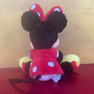 Pu&ograve; includere: Bambola di peluche di Minnie Mouse, vista da dietro, con orecchie e testa di pelo nero, un vestito rosso a pois bianchi e un fiocco abbinato. La bambola ha scarpe gialle e nere e una coda nera.