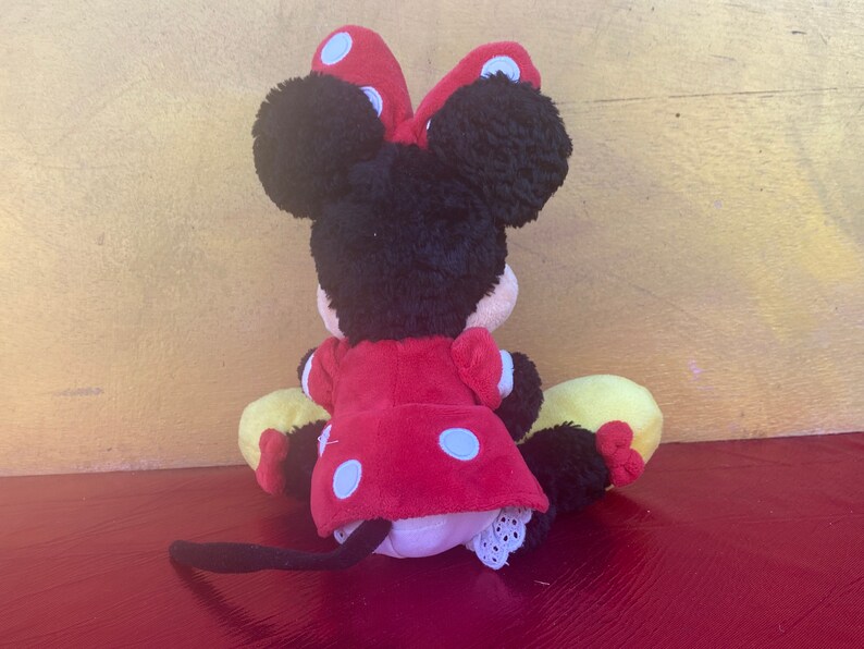 Pu&ograve; includere: Bambola di peluche di Minnie Mouse, vista da dietro, con orecchie e testa di pelo nero, un vestito rosso a pois bianchi e un fiocco abbinato. La bambola ha scarpe gialle e nere e una coda nera.