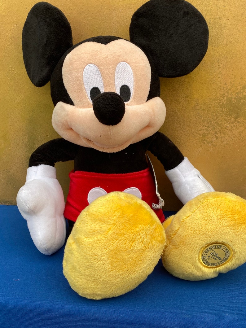 May include: A plush Mickey Mouse doll with black ears, a black nose, and a smiling face. Mickey wears red shorts, white gloves, and yellow shoes. The doll is a Disney Store product.