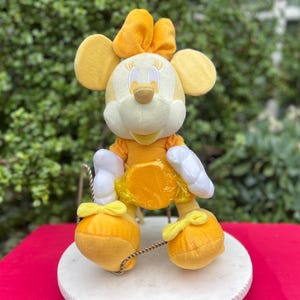Pode incluir: Uma figura de pel&uacute;cia da Minnie Mouse em um traje amarelo e branco, com um la&ccedil;o combinando e orelhas grandes. A figura est&aacute; sentada em uma base circular branca com uma superf&iacute;cie vermelha ao fundo. A figura tem um vestido amarelo e luvas brancas.