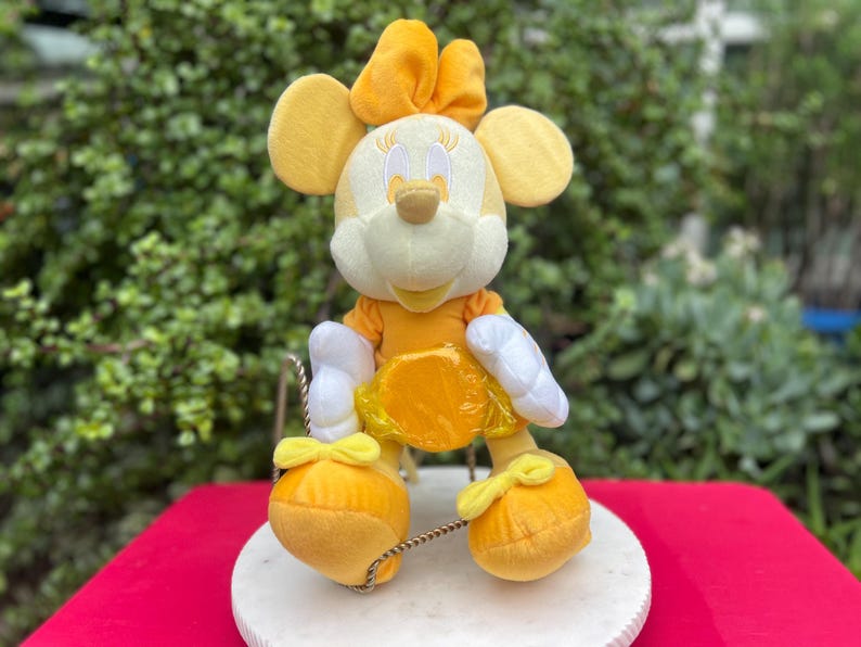 Pode incluir: Uma figura de pel&uacute;cia da Minnie Mouse em um traje amarelo e branco, com um la&ccedil;o combinando e orelhas grandes. A figura est&aacute; sentada em uma base circular branca com uma superf&iacute;cie vermelha ao fundo. A figura tem um vestido amarelo e luvas brancas.