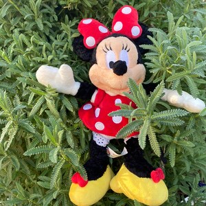 Pu&ograve; includere: Una bambola di peluche di Minnie Mouse con un vestito rosso a pois bianchi, un fiocco abbinato e scarpe gialle. La bambola ha le orecchie nere ed &egrave; circondata da fogliame verde.