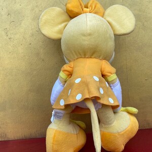 Pode incluir: Um brinquedo de pel&uacute;cia de rato com um vestido amarelo com bolinhas brancas, um la&ccedil;o a condizer e orelhas grandes. O brinquedo tem uma cauda longa e est&aacute; em p&eacute; sobre sapatos amarelos. O brinquedo &eacute; feito de tecido macio e &eacute; um brinquedo infantil.