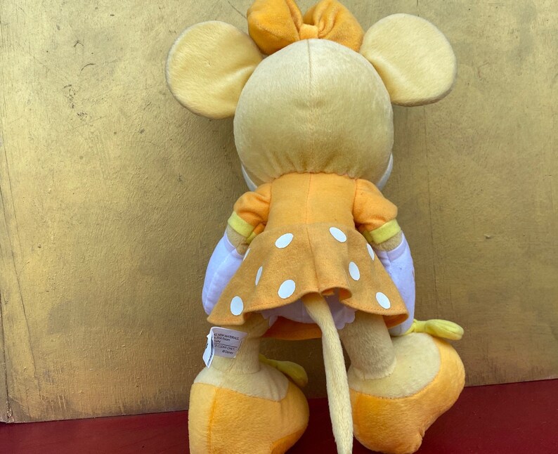 Pode incluir: Um brinquedo de pel&uacute;cia de rato com um vestido amarelo com bolinhas brancas, um la&ccedil;o a condizer e orelhas grandes. O brinquedo tem uma cauda longa e est&aacute; em p&eacute; sobre sapatos amarelos. O brinquedo &eacute; feito de tecido macio e &eacute; um brinquedo infantil.