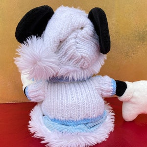 K&ouml;nnte beinhalten: Ein wei&szlig;es Pl&uuml;schspielzeug-H&uuml;ndchen, das eine wei&szlig;e Strickm&uuml;tze mit einem flauschigen Bommel und einen wei&szlig;en Strickpullover mit blauem Besatz tr&auml;gt. Der Hund hat schwarze Ohren und eine wei&szlig;e M&uuml;tze an einer Pfote.