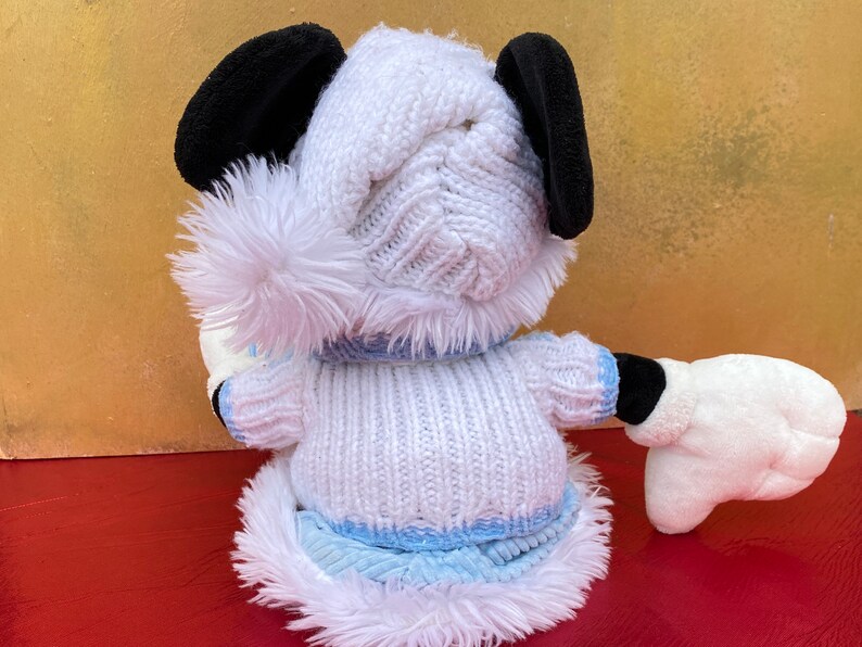 K&ouml;nnte beinhalten: Ein wei&szlig;es Pl&uuml;schspielzeug-H&uuml;ndchen, das eine wei&szlig;e Strickm&uuml;tze mit einem flauschigen Bommel und einen wei&szlig;en Strickpullover mit blauem Besatz tr&auml;gt. Der Hund hat schwarze Ohren und eine wei&szlig;e M&uuml;tze an einer Pfote.
