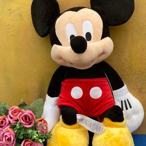 May include: A plush Mickey Mouse doll with black ears, a black head, and a tan face. Mickey wears red shorts with white buttons, white gloves, and yellow shoes. A bouquet of pink roses sits to the left of the doll.