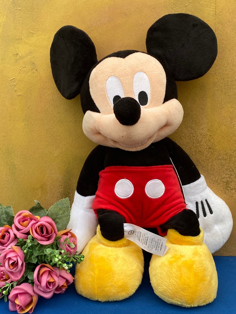 May include: A plush Mickey Mouse doll with black ears, a black head, and a tan face. Mickey wears red shorts with white buttons, white gloves, and yellow shoes. A bouquet of pink roses sits to the left of the doll.