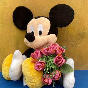 May include: A plush Mickey Mouse doll with black ears, a tan face, and a black body. The doll is holding a bouquet of pink roses with green leaves. The background is a textured gold color.