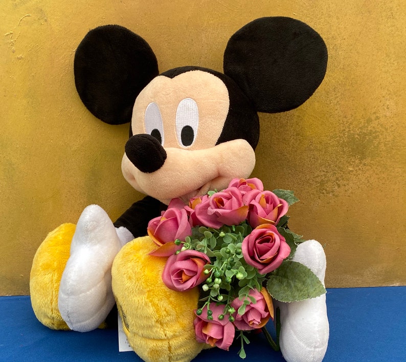 May include: A plush Mickey Mouse doll with black ears, a tan face, and a black body. The doll is holding a bouquet of pink roses with green leaves. The background is a textured gold color.