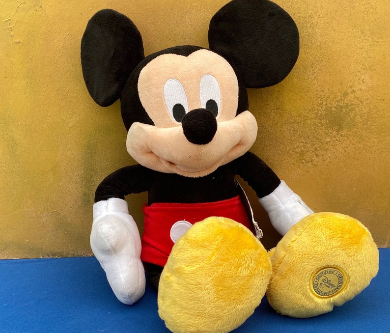 May include: A plush Mickey Mouse toy, featuring black ears, a black body, white gloves, red shorts, and yellow shoes. The toy is sitting on a blue surface with a gold-colored background. A small tag is attached.
