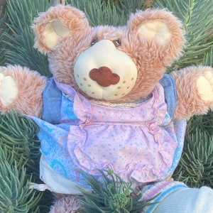 Peut inclure: Un ours en peluche avec une fourrure brun clair, vêtu d'une robe florale bleue et rose avec des bordures à volants. L'ours a un museau crème avec un nez brun, entouré de feuillage vert.