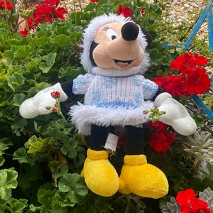 K&ouml;nnte beinhalten: Ein Pl&uuml;schtier von Mickey Mouse, das einen wei&szlig;en Pullover mit blauen Schneeflocken und einem wei&szlig;en Pelzbesatz tr&auml;gt. Das Spielzeug tr&auml;gt gelbe Schuhe und hat eine rote Schleife auf dem Kopf. Das Spielzeug sitzt in einem Bett aus gr&uuml;nen Pflanzen mit roten Blumen.