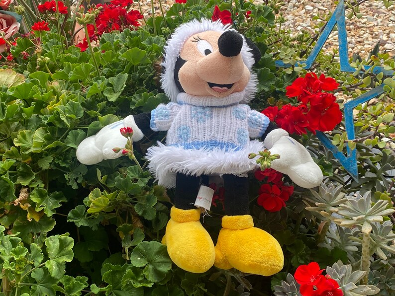 K&ouml;nnte beinhalten: Ein Pl&uuml;schtier von Mickey Mouse, das einen wei&szlig;en Pullover mit blauen Schneeflocken und einem wei&szlig;en Pelzbesatz tr&auml;gt. Das Spielzeug tr&auml;gt gelbe Schuhe und hat eine rote Schleife auf dem Kopf. Das Spielzeug sitzt in einem Bett aus gr&uuml;nen Pflanzen mit roten Blumen.