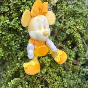 Pode incluir: Uma boneca de pel&uacute;cia da Minnie Mouse sentada. A boneca apresenta um la&ccedil;o, vestido e sapatos amarelos, luvas brancas e um rosto creme. O fundo &eacute; folhagem verde.