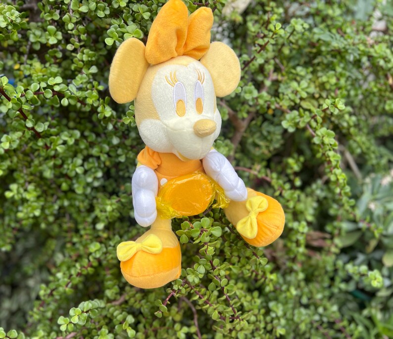 Pode incluir: Uma boneca de pel&uacute;cia da Minnie Mouse sentada. A boneca apresenta um la&ccedil;o, vestido e sapatos amarelos, luvas brancas e um rosto creme. O fundo &eacute; folhagem verde.