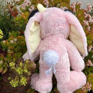 May include: A pink plush bunny rabbit with yellow and white checkered ears and a white fluffy tail. The bunny is sitting on a bed of green plants with small white flowers.