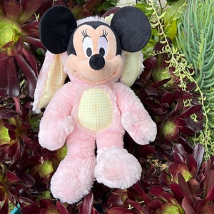 May include: Pink plush bunny costume with yellow and white checkered accents. The costume features a Mickey Mouse head with black ears and a black nose. The costume is sitting on a bed of green and brown plants.