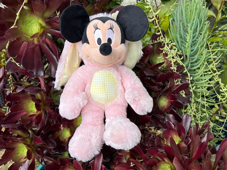May include: Pink plush bunny costume with yellow and white checkered accents. The costume features a Mickey Mouse head with black ears and a black nose. The costume is sitting on a bed of green and brown plants.