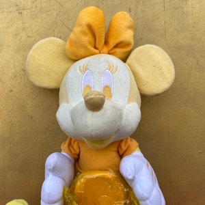 Pode incluir: Uma boneca de pel&uacute;cia da Minnie Mouse em tons de amarelo e branco. A boneca tem orelhas grandes e redondas, um la&ccedil;o na cabe&ccedil;a e um vestido amarelo. A boneca &eacute; macia e fofa, perfeita para crian&ccedil;as.