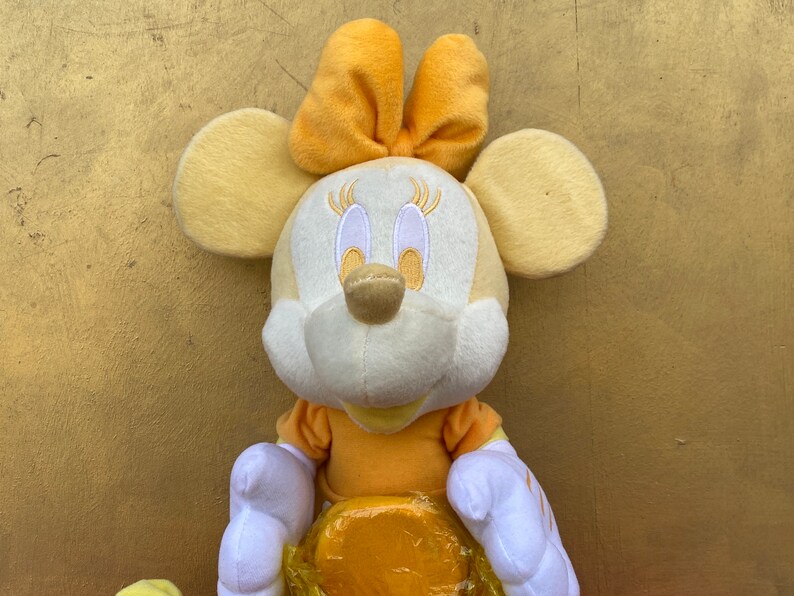 Pode incluir: Uma boneca de pel&uacute;cia da Minnie Mouse em tons de amarelo e branco. A boneca tem orelhas grandes e redondas, um la&ccedil;o na cabe&ccedil;a e um vestido amarelo. A boneca &eacute; macia e fofa, perfeita para crian&ccedil;as.