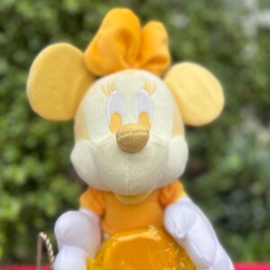 Pode incluir: Um brinquedo de pel&uacute;cia de um personagem com orelhas amarelas, um la&ccedil;o amarelo e um rosto amarelo e branco. O personagem est&aacute; vestindo um vestido amarelo e luvas brancas. O fundo &eacute; um verde desfocado.