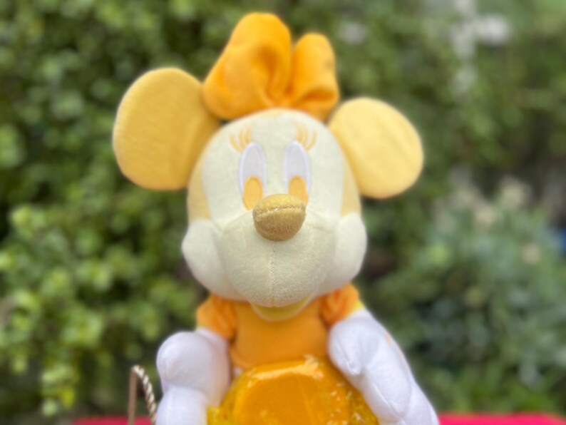 Pode incluir: Um brinquedo de pel&uacute;cia de um personagem com orelhas amarelas, um la&ccedil;o amarelo e um rosto amarelo e branco. O personagem est&aacute; vestindo um vestido amarelo e luvas brancas. O fundo &eacute; um verde desfocado.